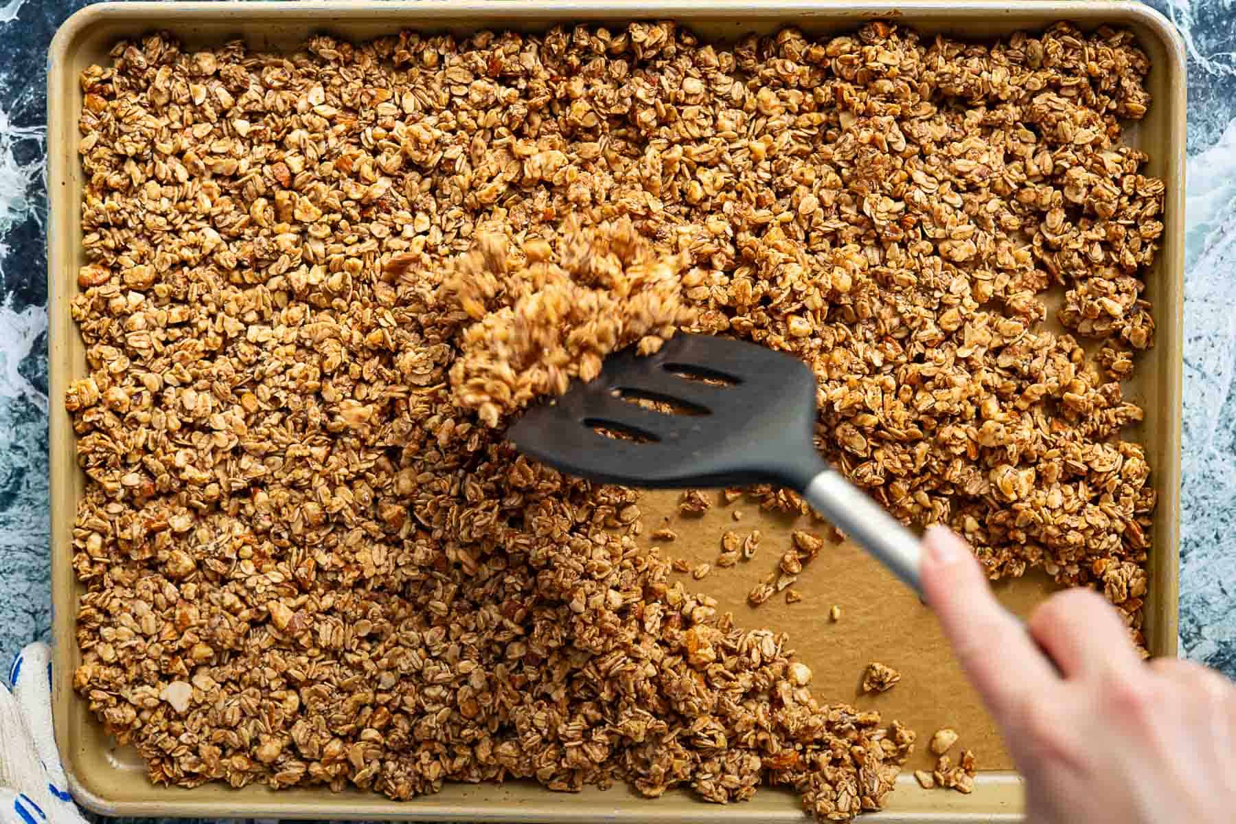 A hand using a black spatula to stir golden-brown sourdough discard granola on a baking sheet, spreading the mixture evenly across the surface.