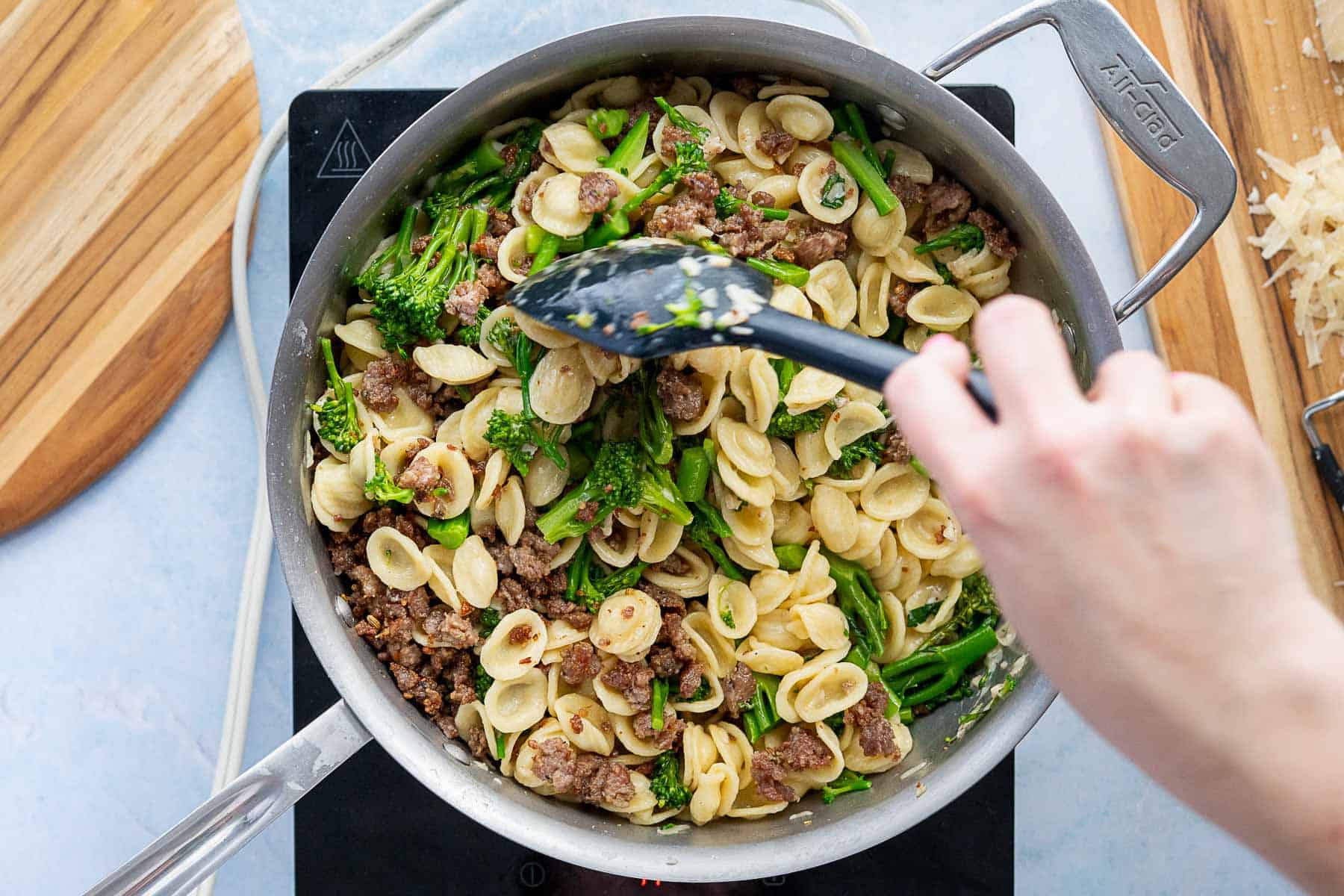A hand stirs broccolini pasta with orecchiette and sausage in a large pan on a stovetop. Grated cheese sits on a nearby cutting board.