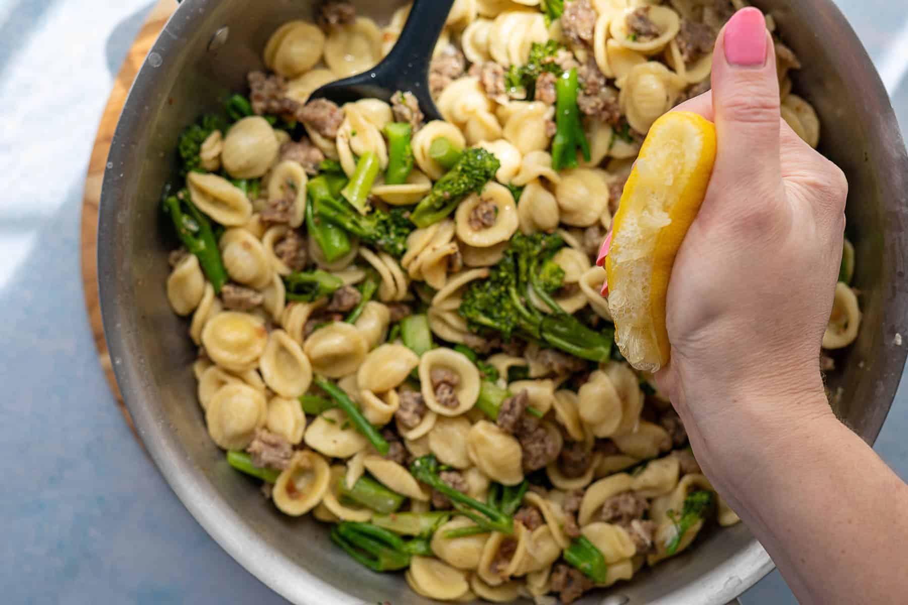 A hand squeezes a lemon over a pan of broccolini pasta with orecchiette and sausage. A black serving spoon rests in the dish, and the person’s pink nail polish adds a splash of color.