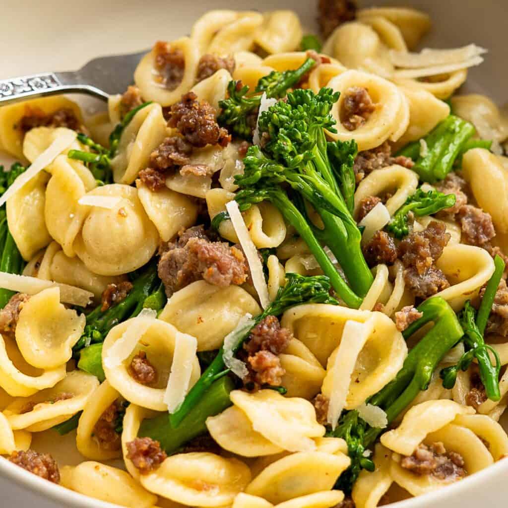 A close-up of broccolini pasta features orecchiette mixed with crumbled sausage, vibrant green broccolini, and a sprinkle of grated parmesan cheese. A fork is partially visible on the side.