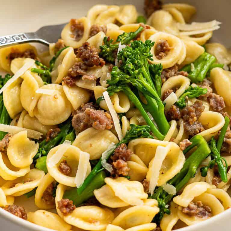 A close-up of broccolini pasta features orecchiette mixed with crumbled sausage, vibrant green broccolini, and a sprinkle of grated parmesan cheese. A fork is partially visible on the side.