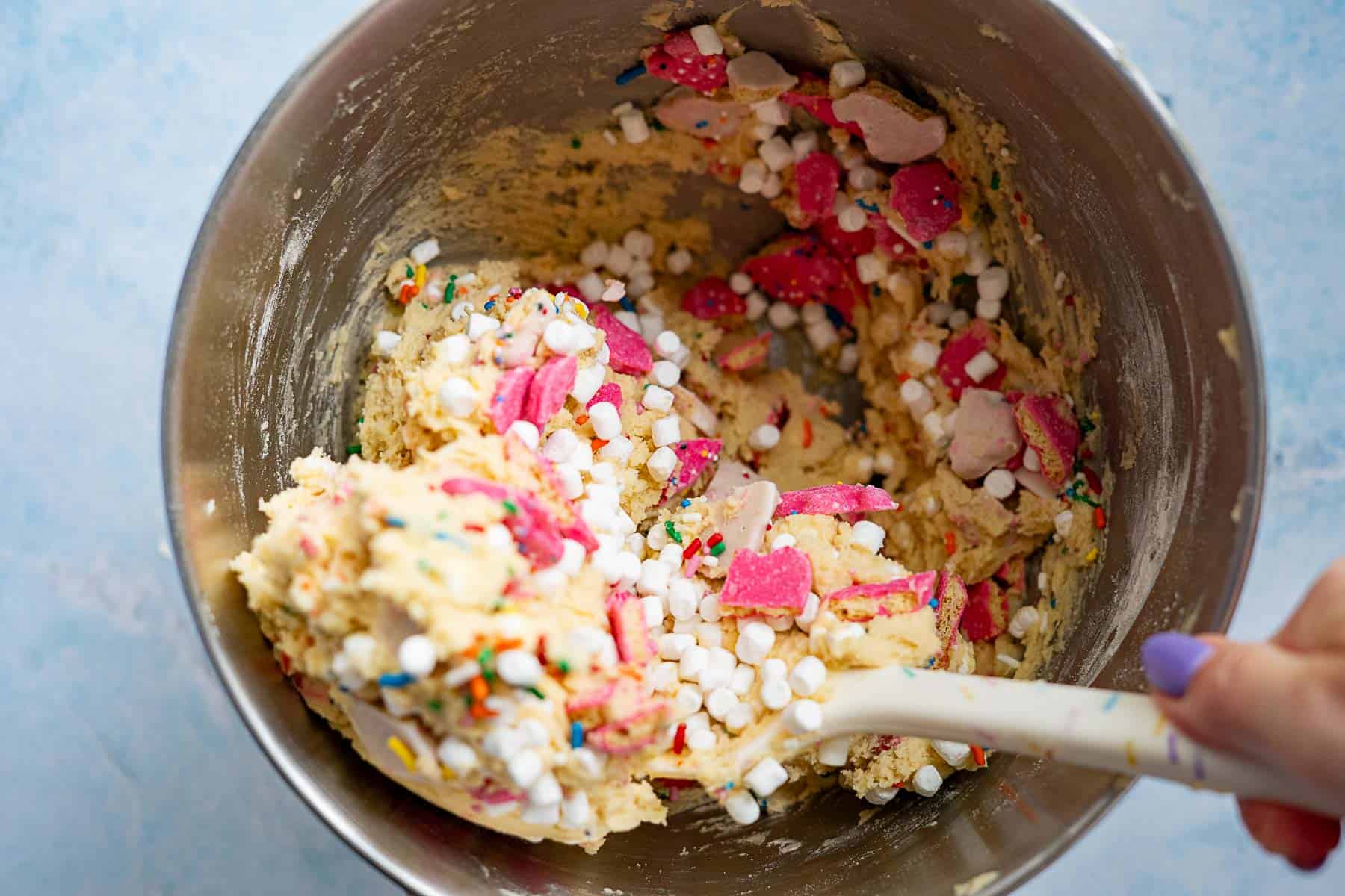 A mixing bowl filled with cookie dough containing colorful sprinkles, circus animal cookies, and small white marshmallows. A hand is stirring the mixture with a white spatula.