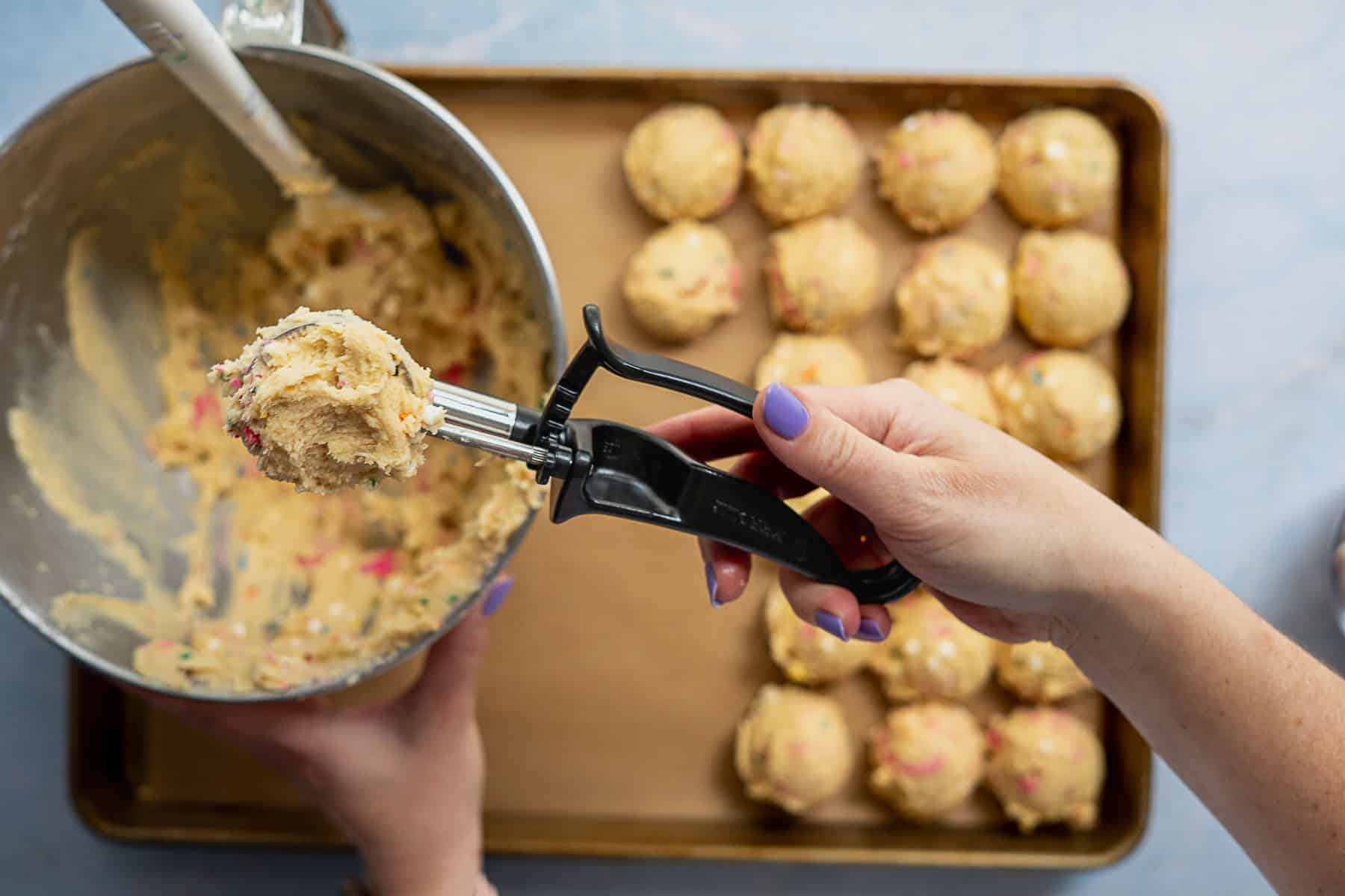 A person scoops circus animal cookie dough with a cookie scoop over a mixing bowl, while a baking tray with evenly spaced dough balls sits on a parchment-lined baking sheet in the background.