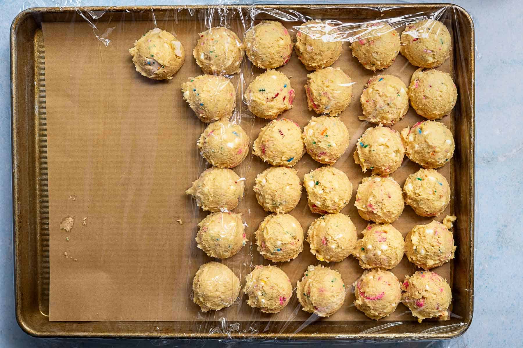 Scoops of unbaked cookie dough with colorful sprinkles and bits of circus animal cookies are arranged in rows on a parchment-lined baking sheet, partially covered with plastic wrap.