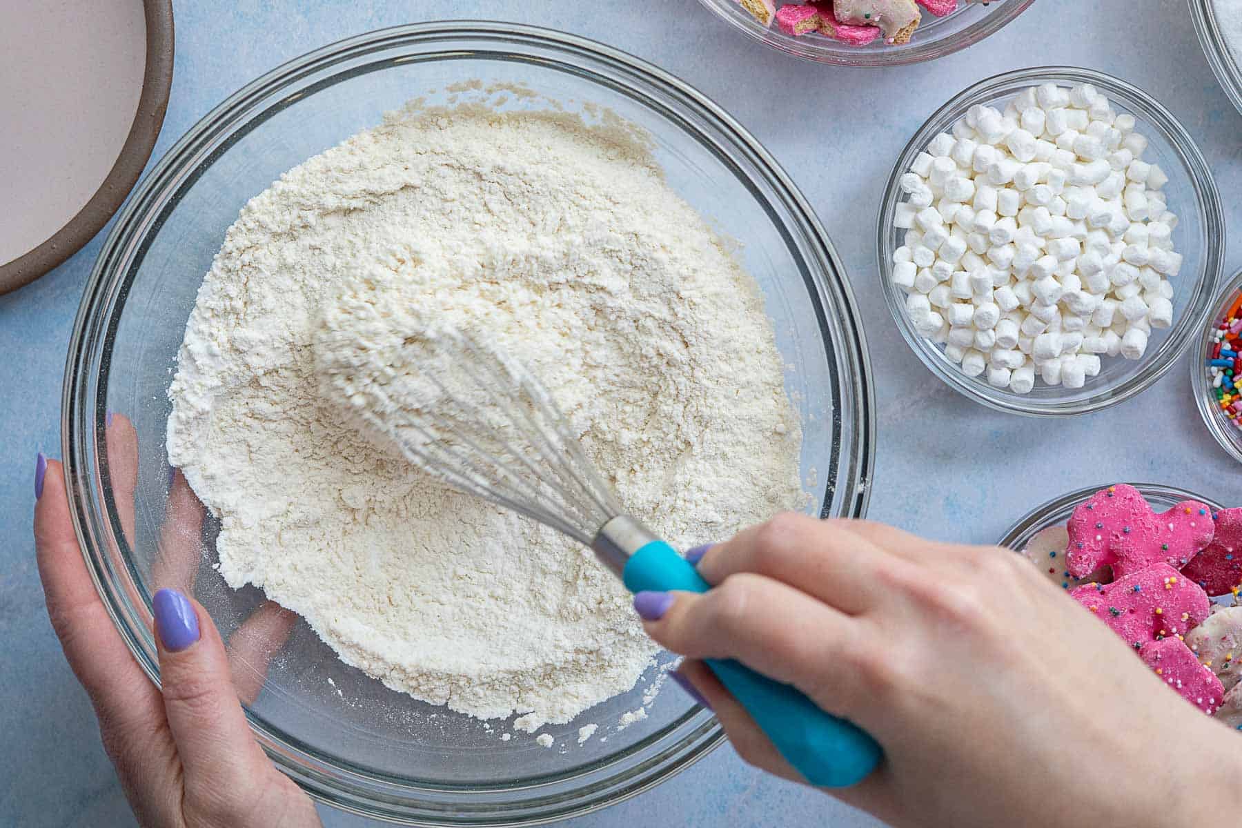 A person with purple-painted nails uses a whisk to mix flour in a glass bowl. Surrounding bowls contain mini marshmallows, colorful sprinkles, and circus animal cookies for a fun and playful baking session.