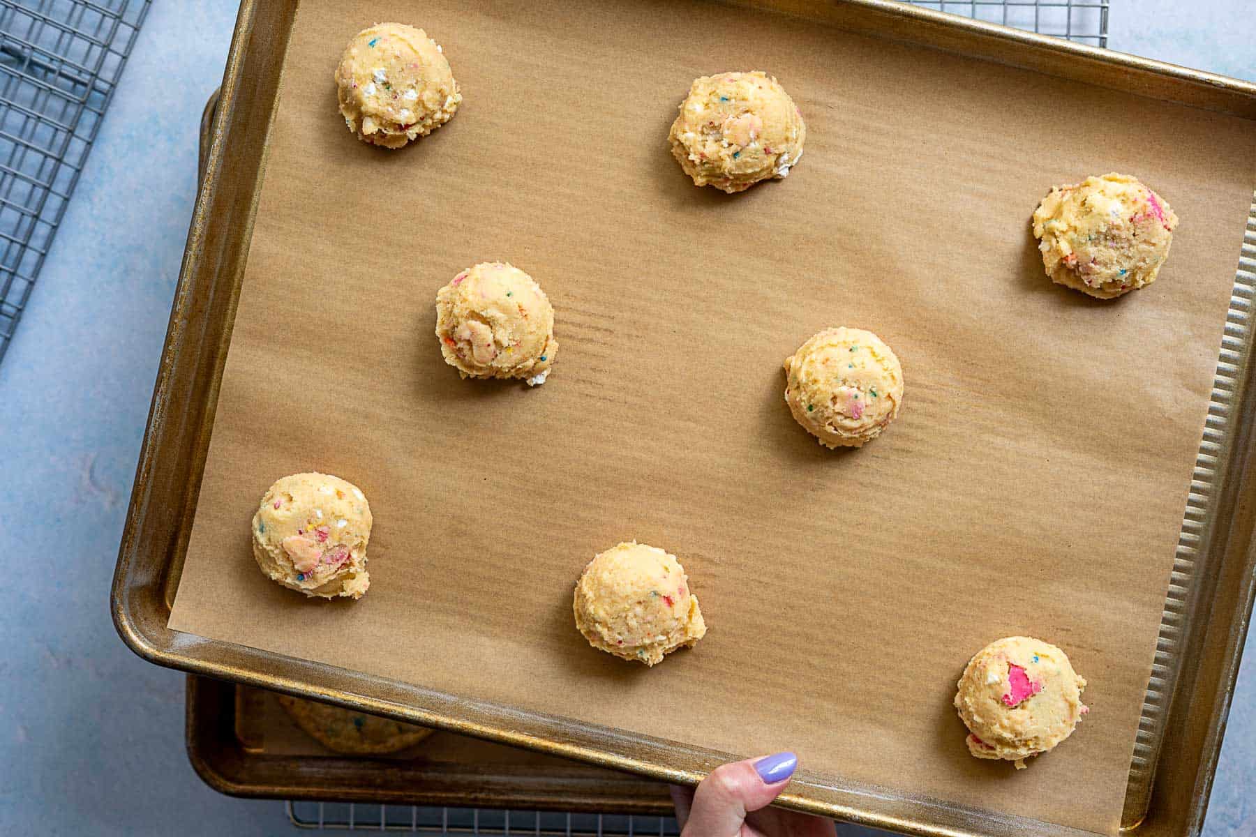 A hand holds a baking sheet lined with parchment paper, topped with eight evenly spaced scoops of circus animal cookie dough. Cooling racks and another baking sheet are partially visible in the background.