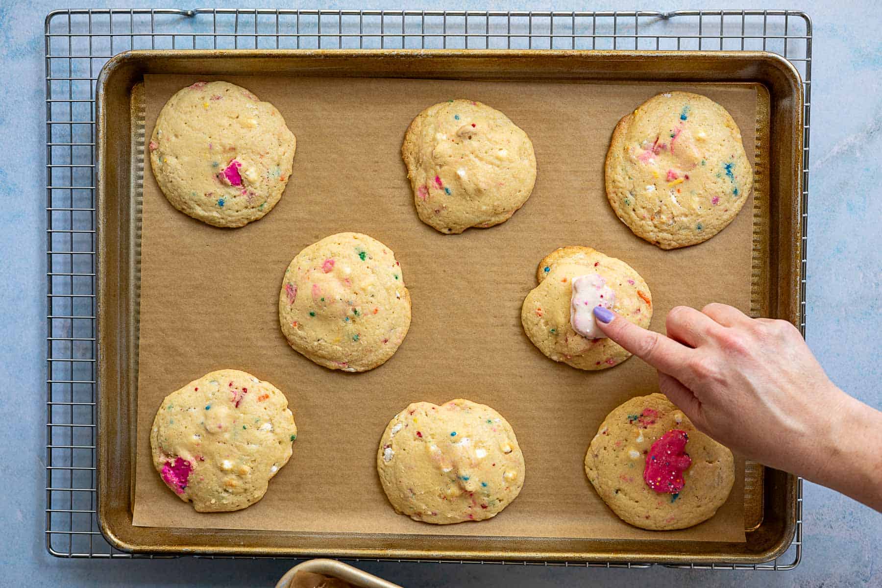 A hand adds a circus animal cookies on top of a round cookie, creating circus animal cookies on a baking sheet lined with parchment paper. Seven other sprinkle-topped cookies surround it, and the tray rests on a cooling rack.