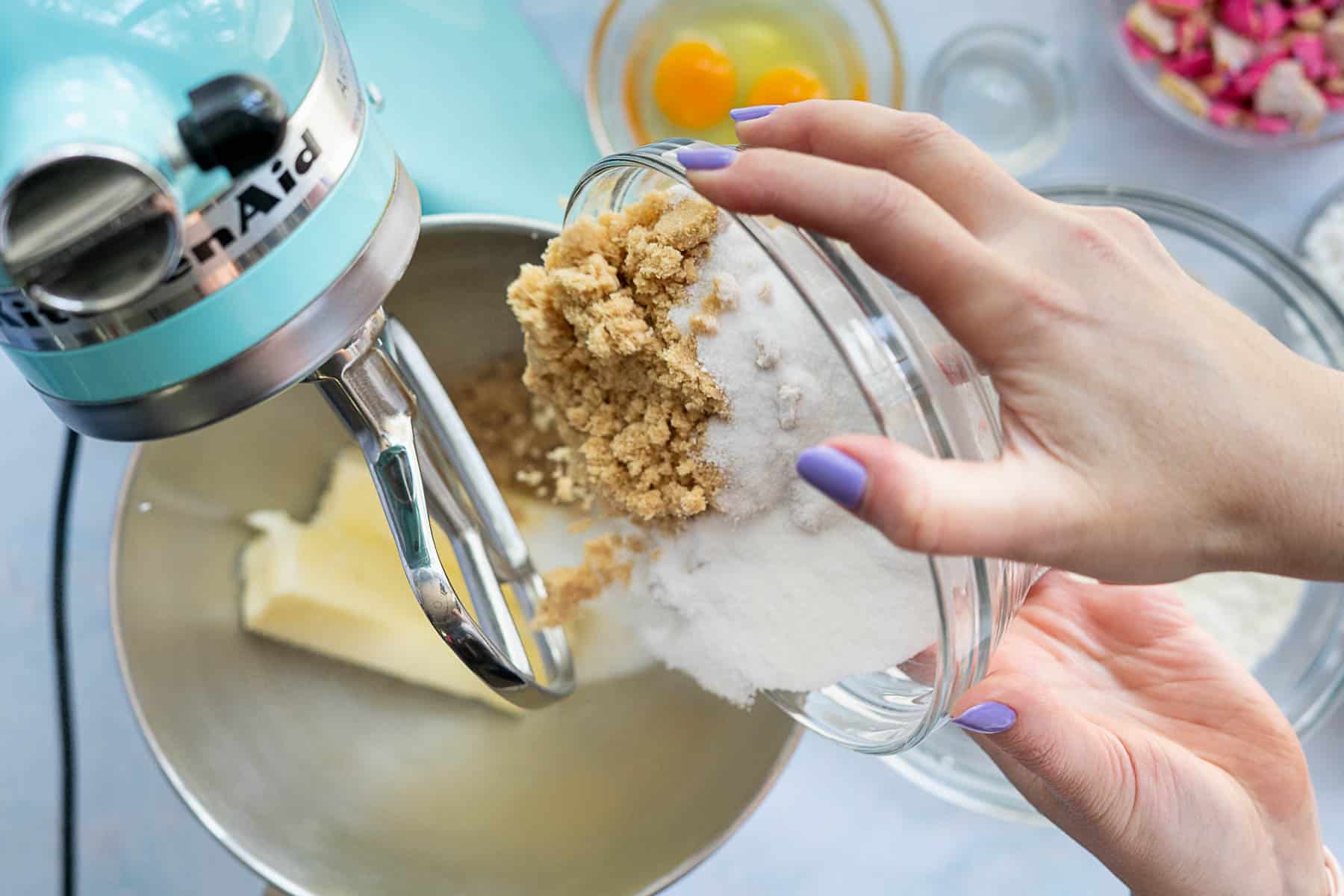 A person with lavender-painted nails pours brown and white sugar from a glass bowl into a stand mixer with butter, preparing ingredients for circus animal cookies. Other baking items and eggs are visible in the background.