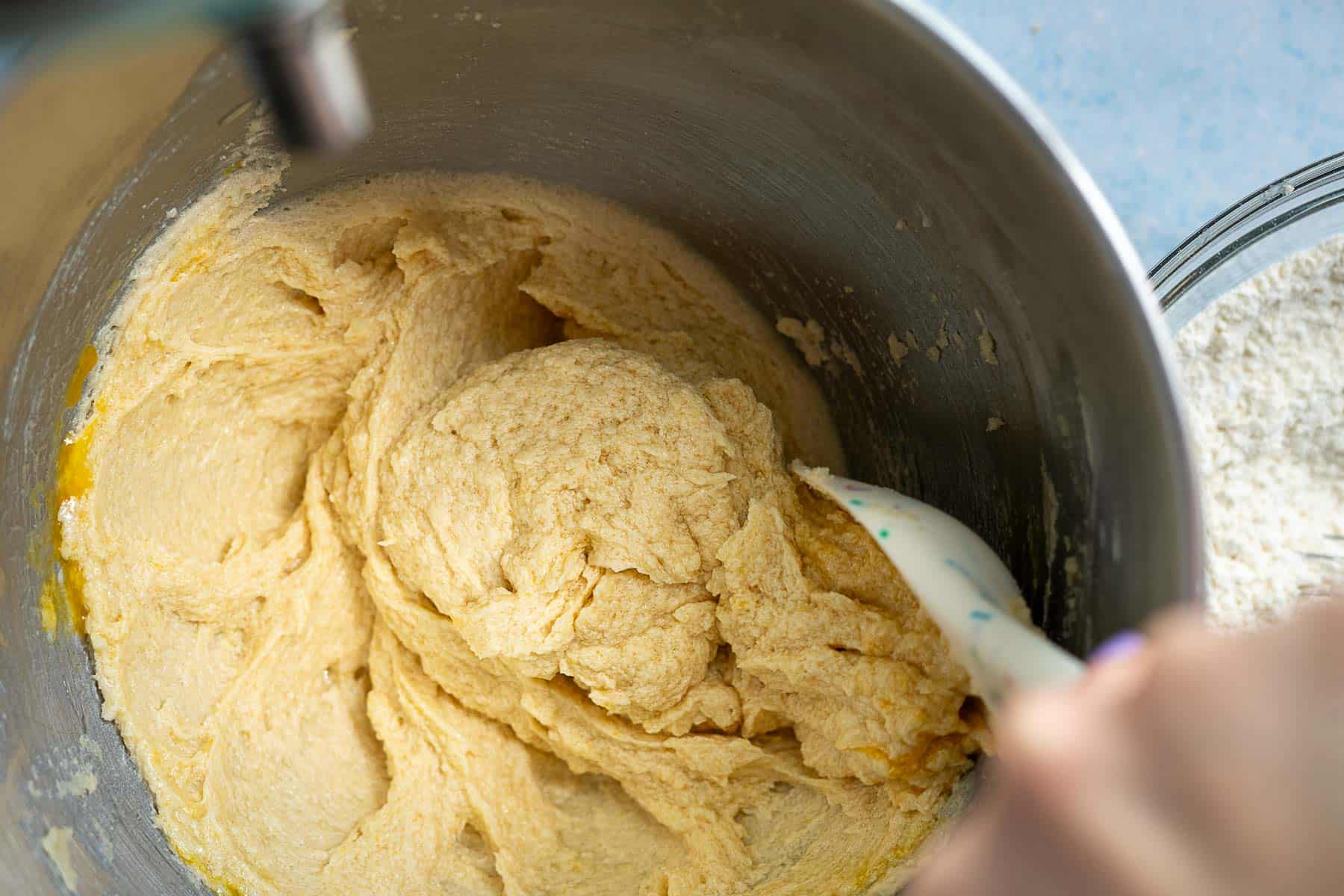 A close-up of cookie dough being mixed in a metal bowl with a spatula. Some dry flour is visible on the right edge, ready to be incorporated along with colorful circus animal cookies for extra fun.