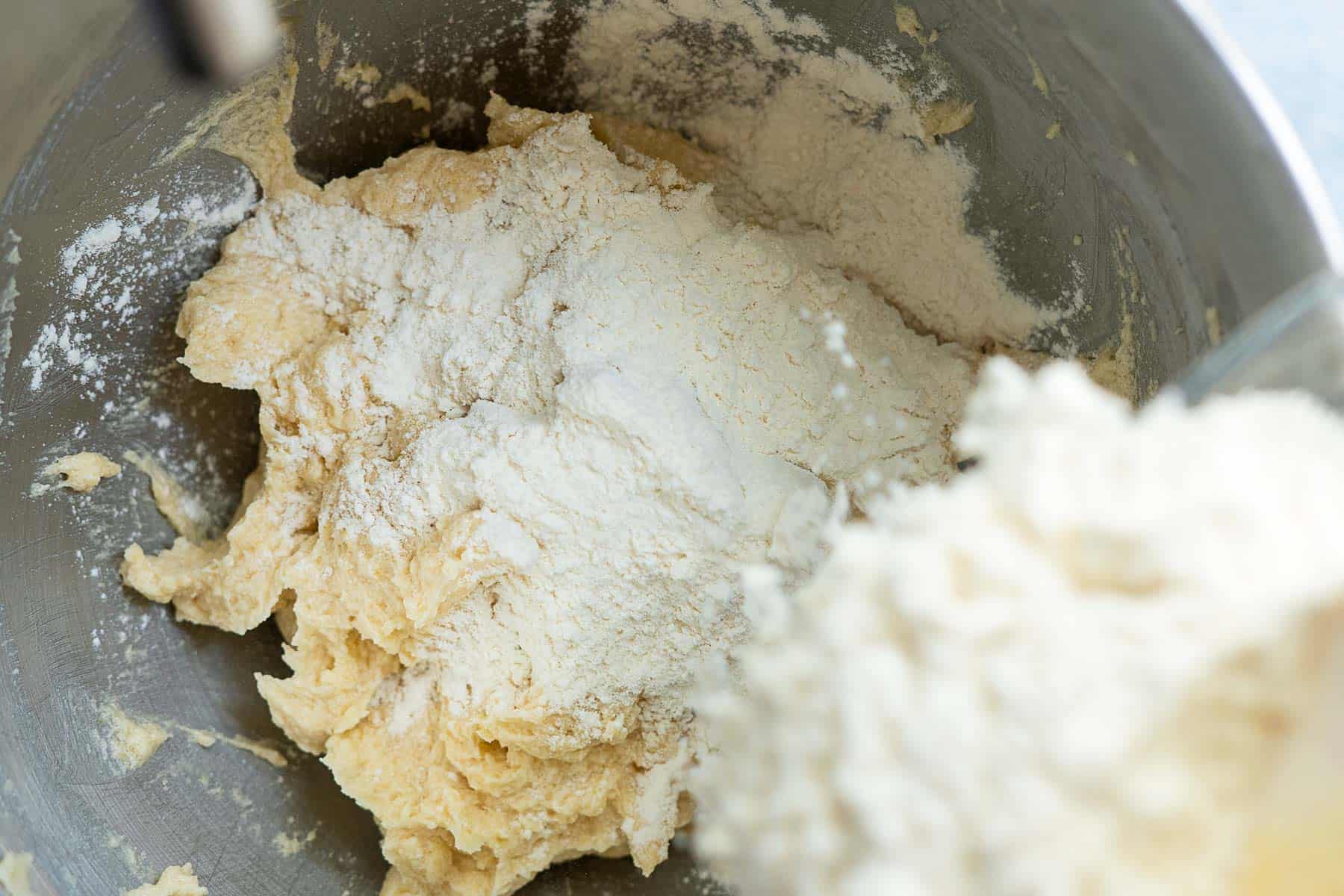 A close-up of flour being added to a mixing bowl of yellow cookie dough, with circus animal cookies ready nearby and the ingredients partially blended.