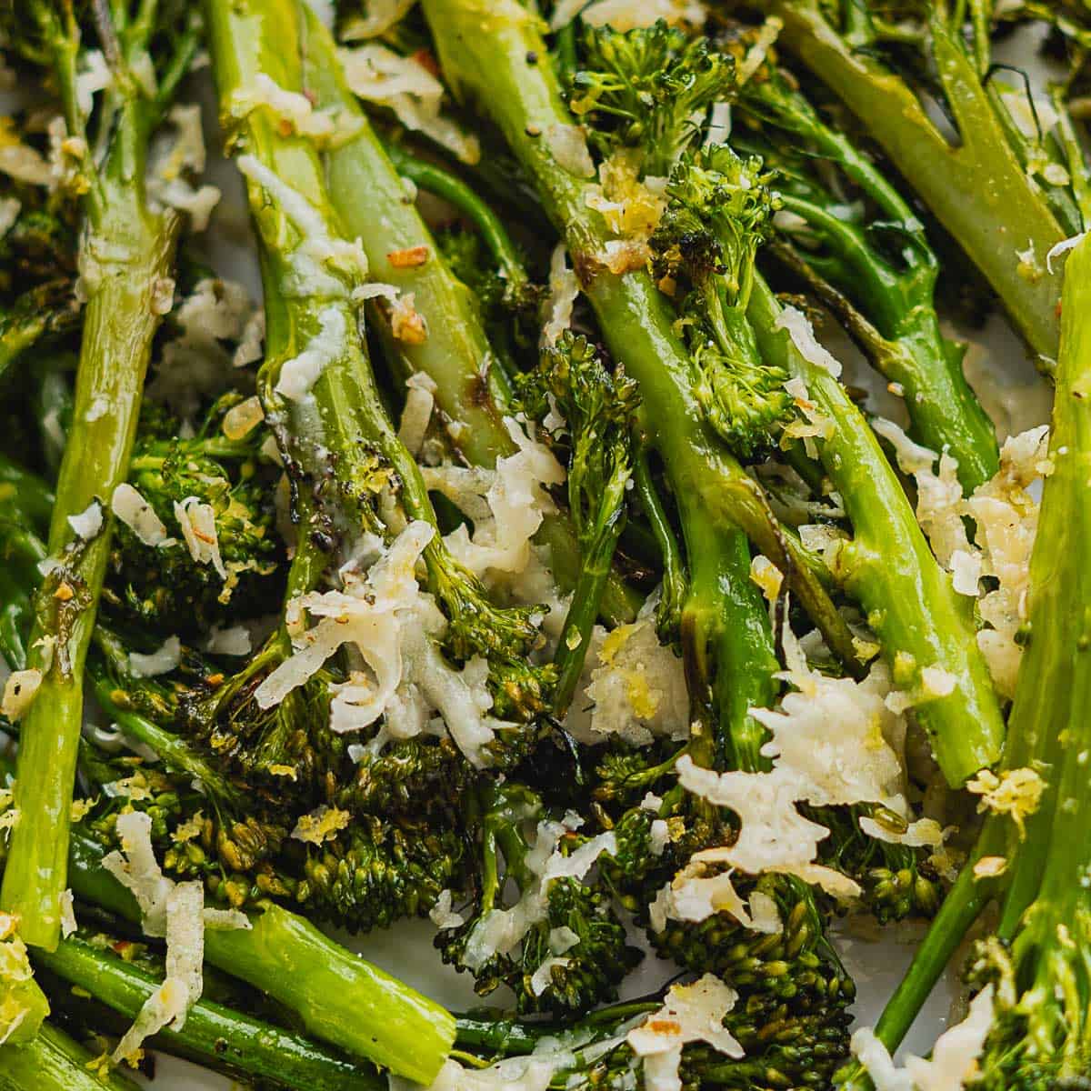 Close-up of roasted broccolini topped with melted grated cheese, highlighting crispy edges and a golden, slightly browned texture. The roasted broccolini stems and florets appear tender, well-seasoned, and perfectly cooked.