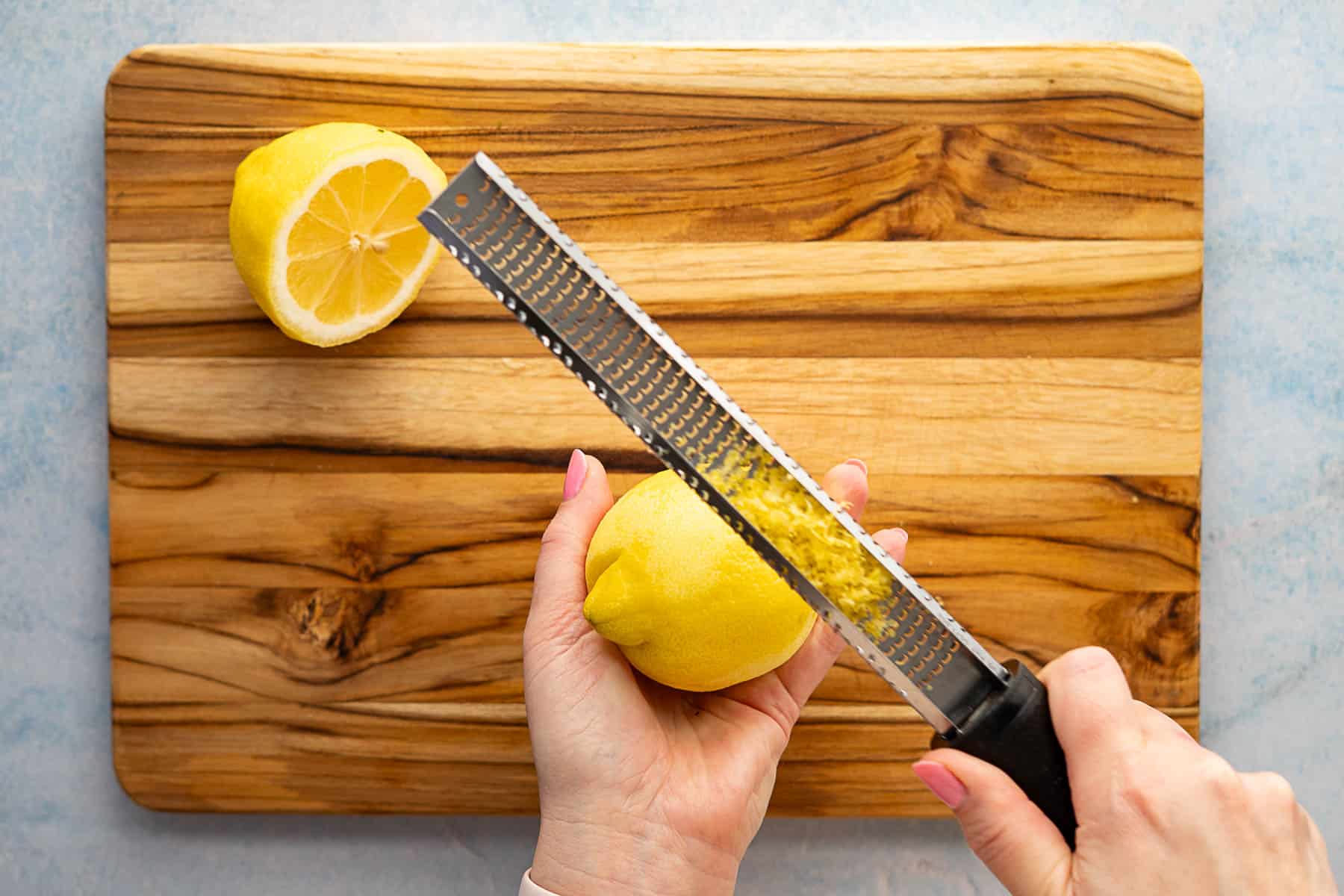 A person zesting a lemon with a grater over a wooden cutting board, preparing fresh zest to sprinkle over roasted broccolini, with a halved lemon resting on the board.