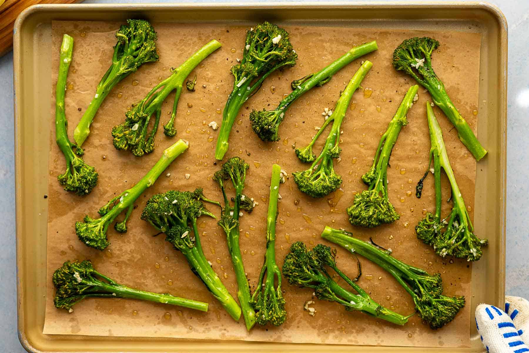 A baking tray lined with parchment paper holds several pieces of roasted broccolini, seasoned with chopped garlic and olive oil, ready for roasting.