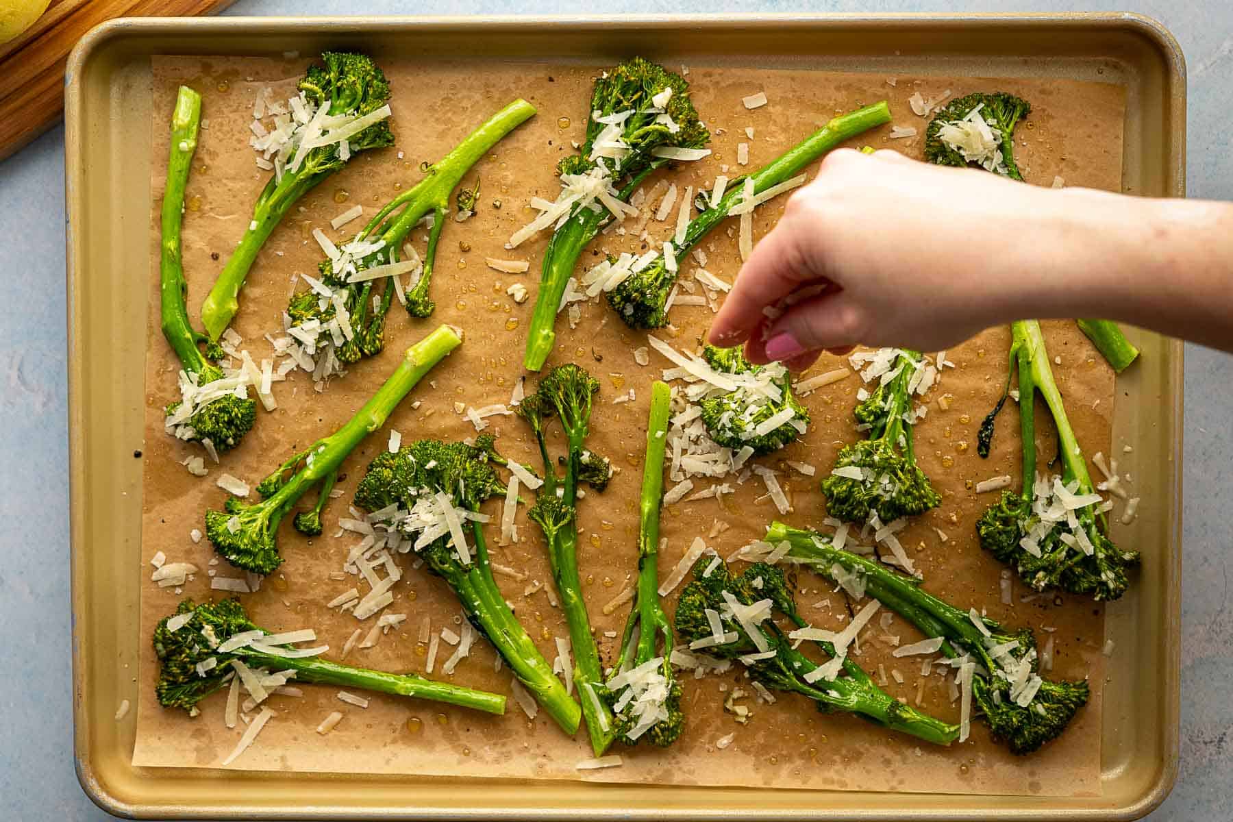 A hand sprinkles grated cheese over roasted broccolini arranged on a parchment-lined baking sheet, ready to be enjoyed.