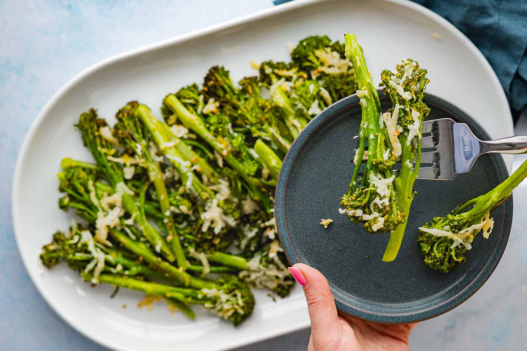 A hand holds a small gray plate with roasted broccolini and melted cheese, while a fork lifts a few stalks. More roasted broccolini with cheese is arranged on a large white platter in the background.