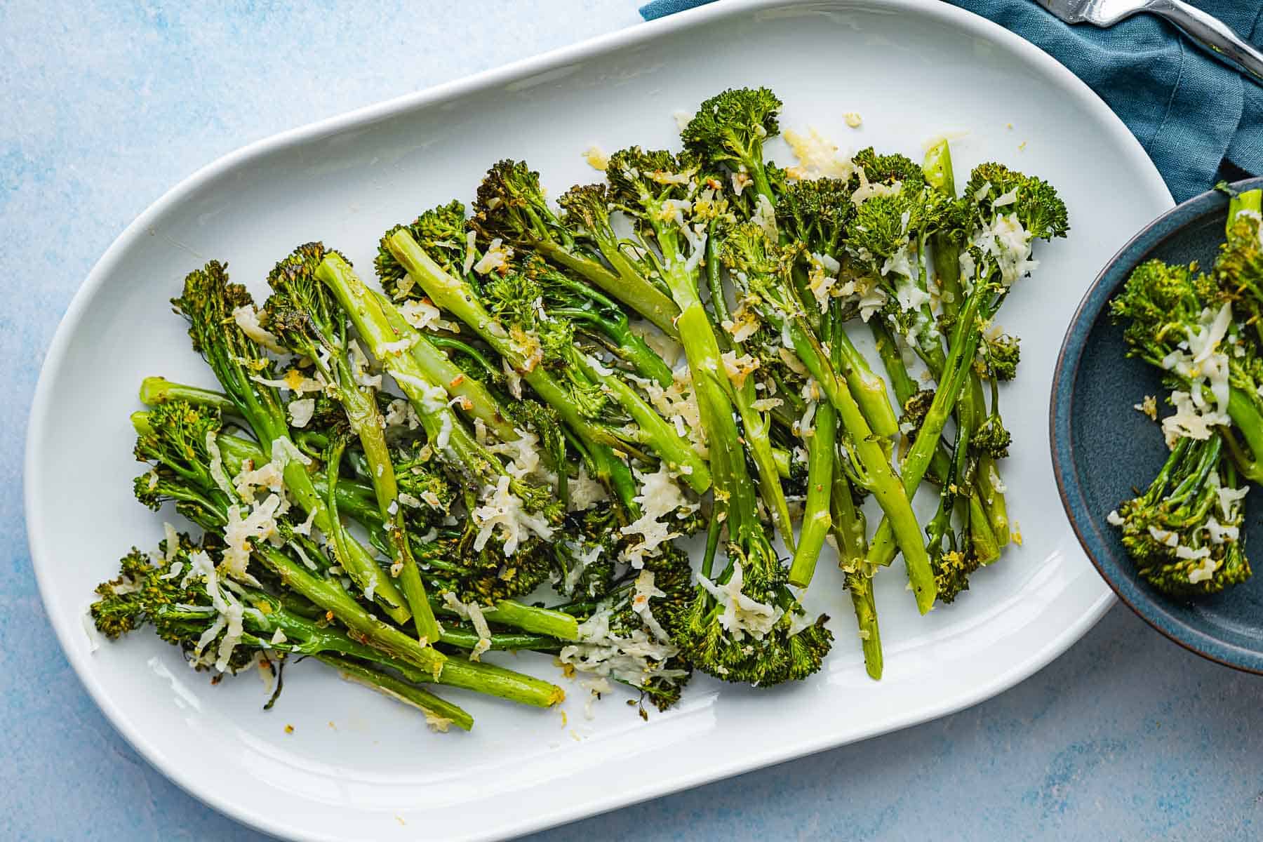 A white platter with roasted broccolini, topped with grated cheese and seasonings, sits next to a small bowl of more roasted broccolini on a light blue surface.