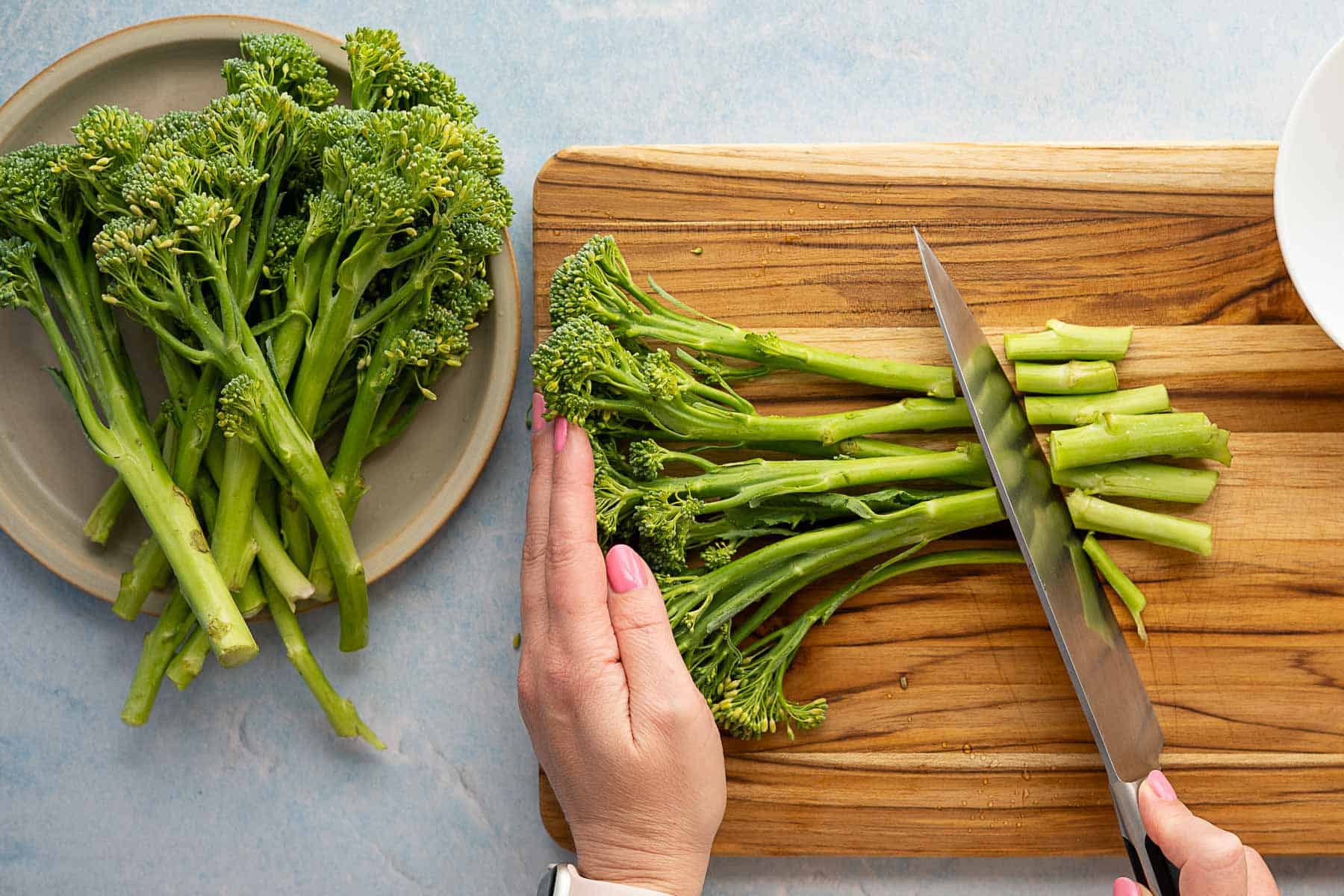 A person slices broccolini on a wooden cutting board with a knife, while a plate with more roasted broccolini sits nearby on a light blue surface.