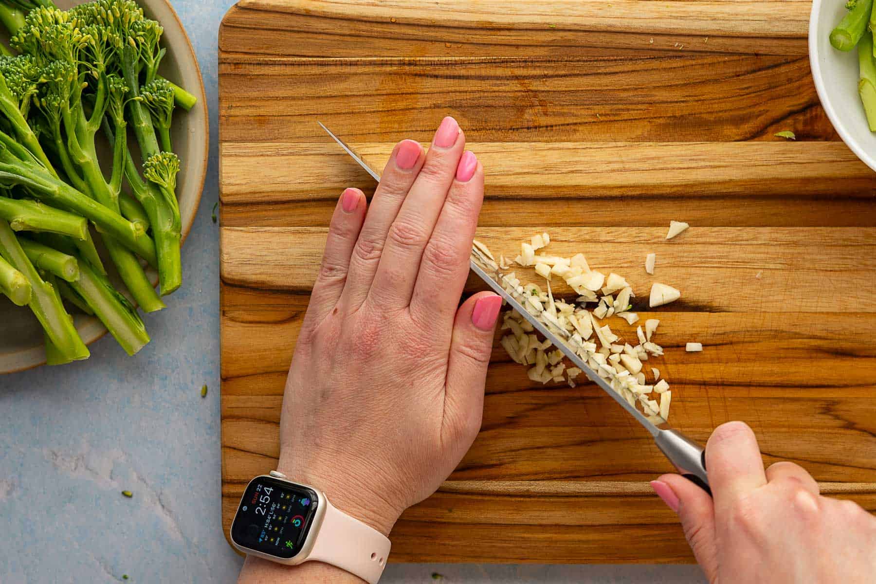 A person wearing a smartwatch chops garlic on a wooden cutting board, with roasted broccolini on a plate nearby.