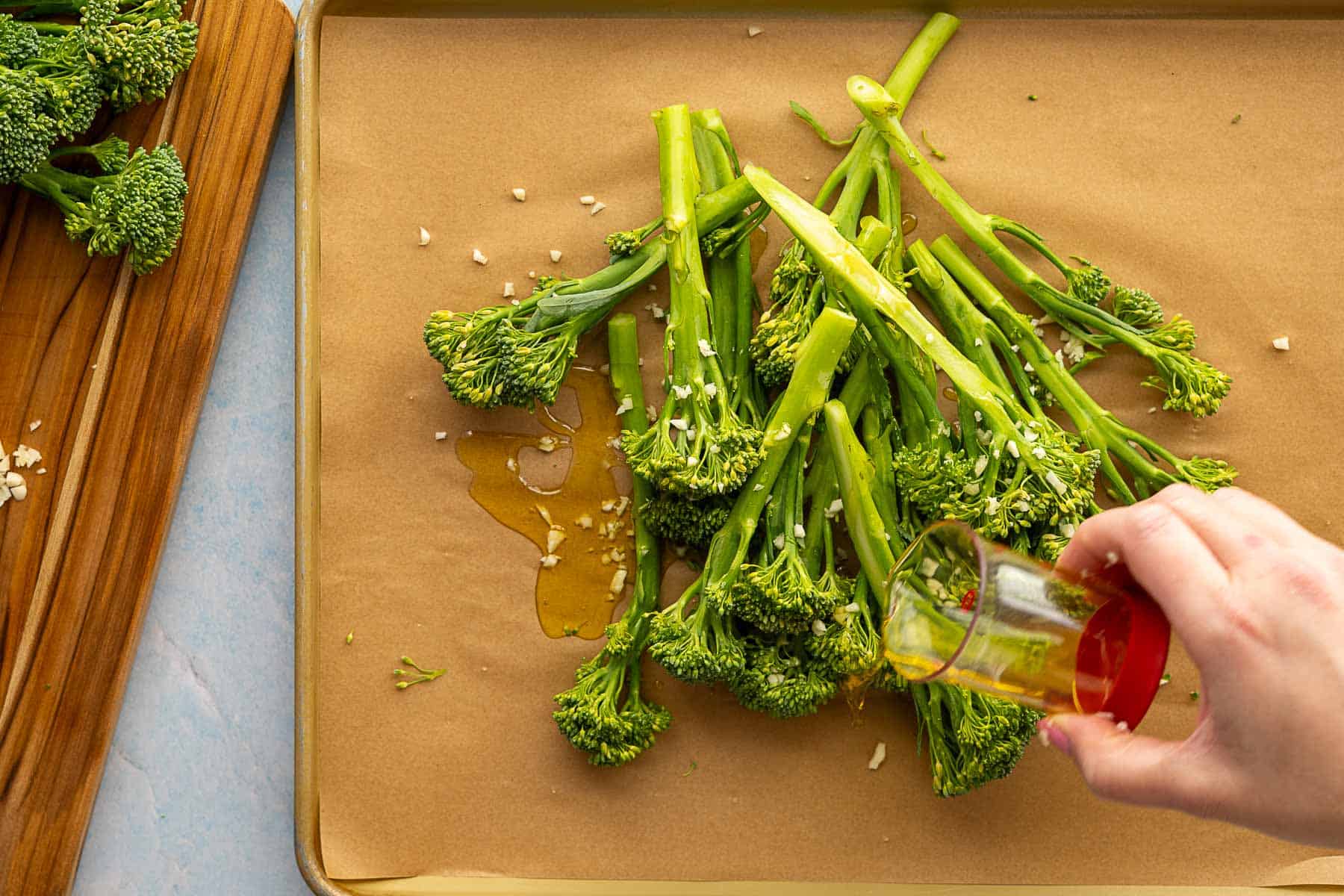 A hand pours olive oil over a pile of fresh broccolini and minced garlic on a parchment-lined baking sheet, preparing delicious roasted broccolini for the oven.
