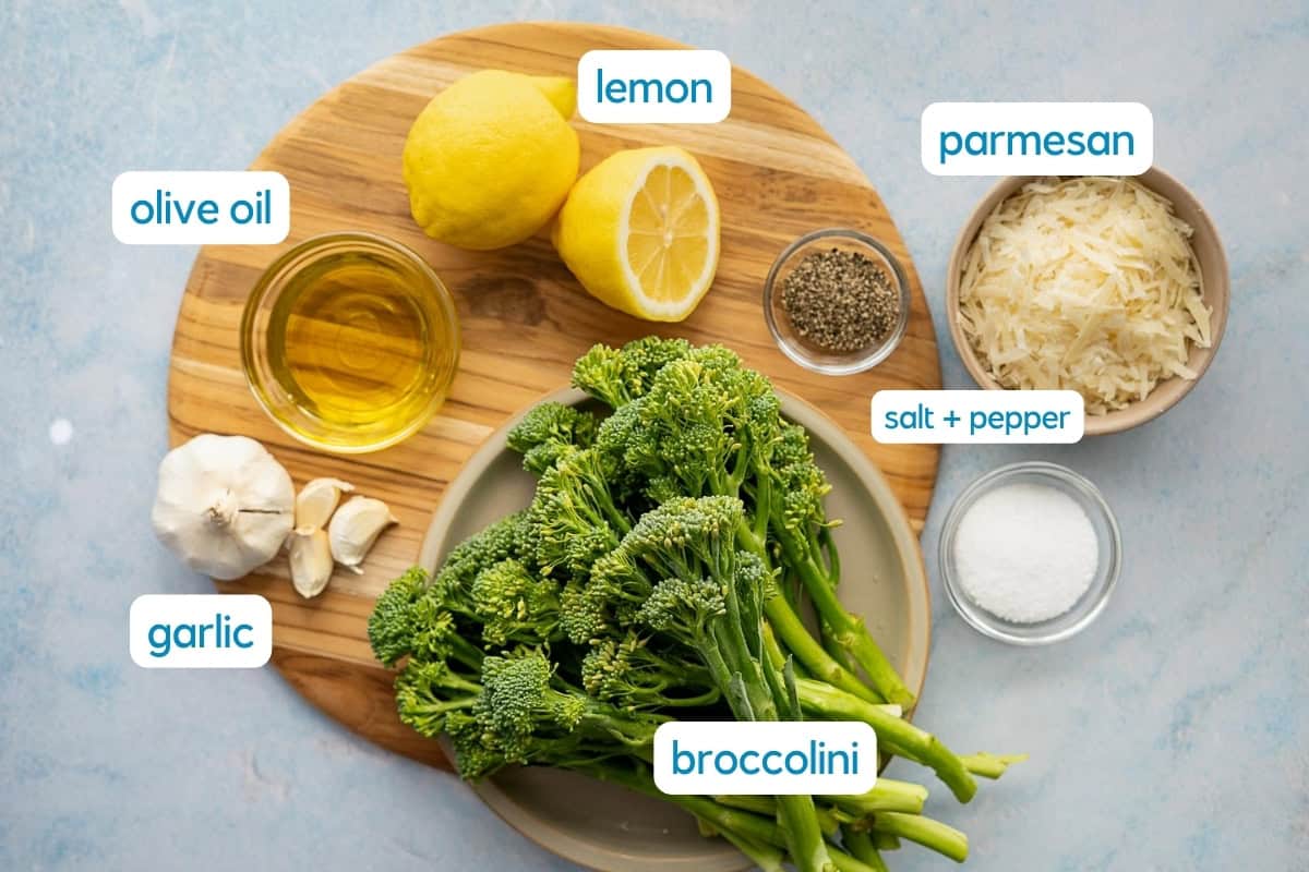 A cutting board with lemons, olive oil, garlic, salt, pepper, parmesan cheese, and a plate of roasted broccolini, each labeled with text.