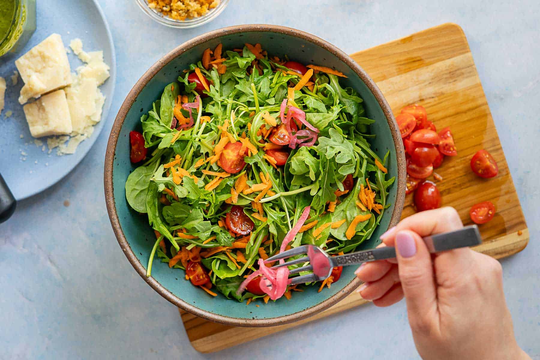 A hand holds a fork with pickled onions over a bowl of salad containing arugula, shredded carrots, cherry tomatoes, and red onions. A plate with cheese and a cutting board with chopped tomatoes are nearby.