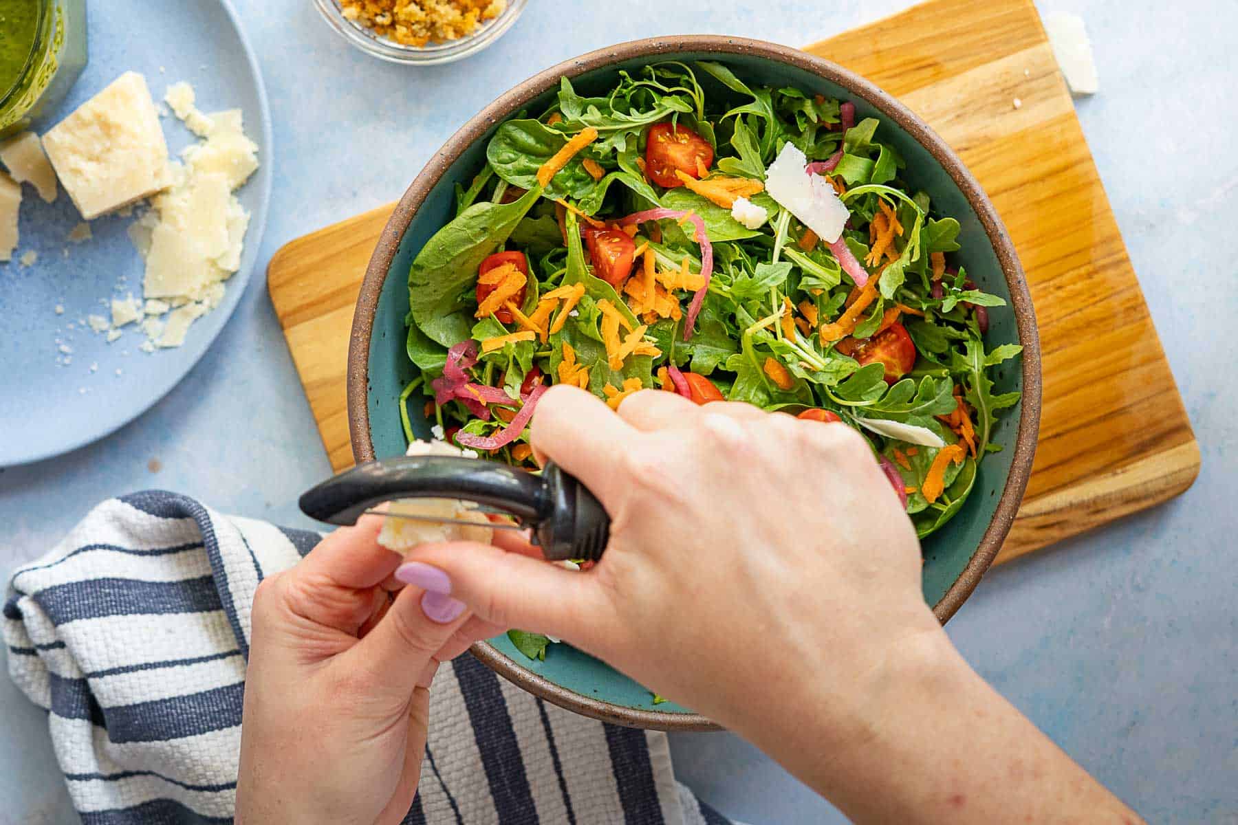 A person uses a peeler to shave cheese over a fresh salad with greens, tomatoes, carrots, and onions in a blue bowl on a wooden board.