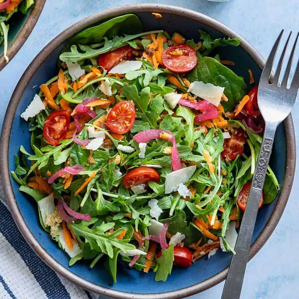 A bowl of mixed green spinach and arugula salad with cherry tomatoes, shredded carrots, shaved cheese, and red onions, with a fork beside it.