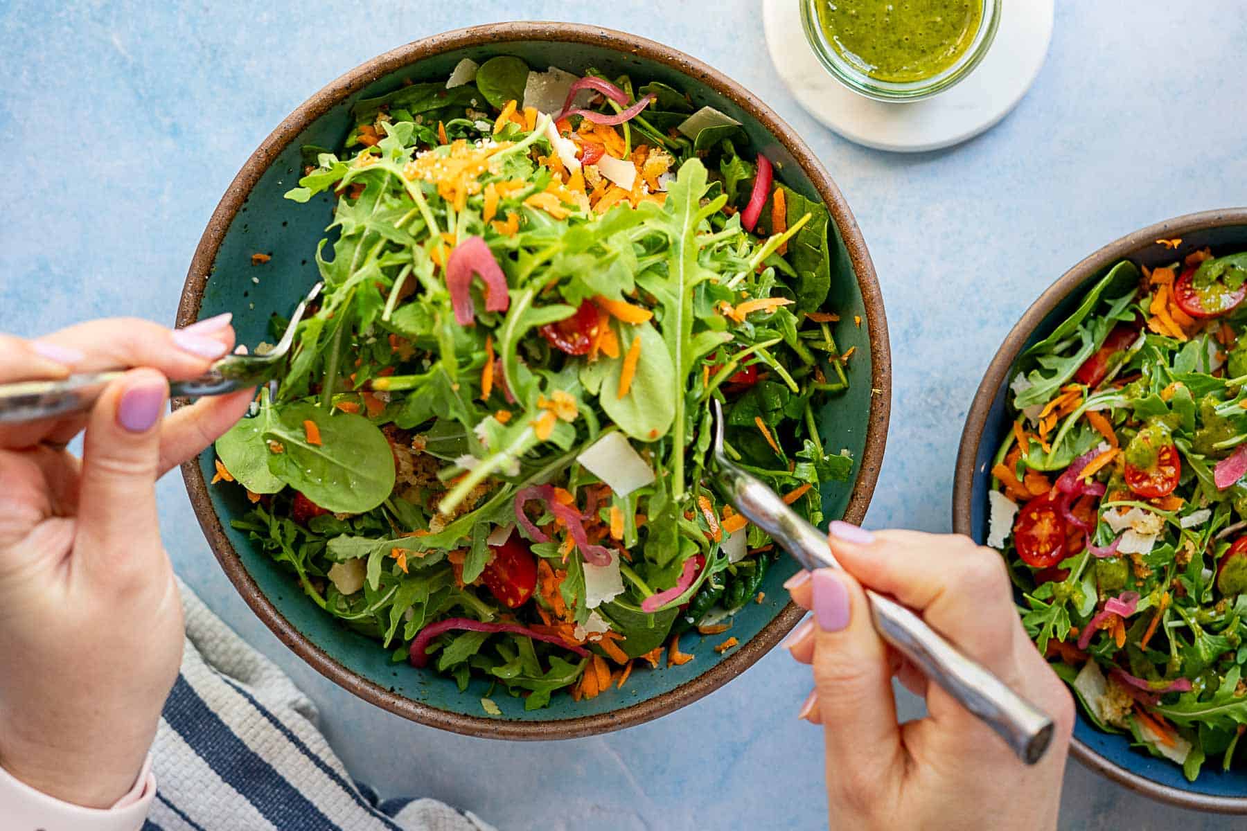 A person with light purple nail polish holds a fork and knife, preparing to eat a fresh spinach salad with arugula, cherry tomatoes, shredded carrots, and red onions, with a jar of green dressing nearby.