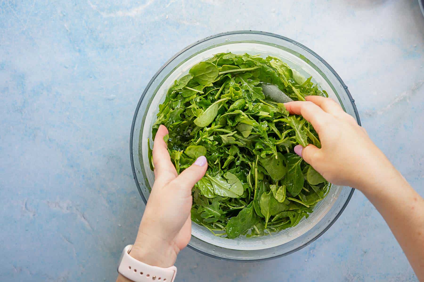 A person wearing a smartwatch tosses fresh green leafy salad in a glass bowl on a light blue countertop.