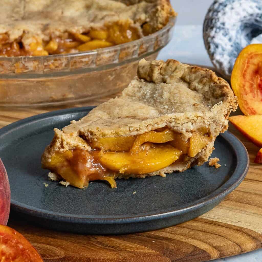 A slice of peach pie with golden crust and juicy peach filling sits on a dark plate, with the rest of the pie and fresh peach slices in the background on a wooden surface.
