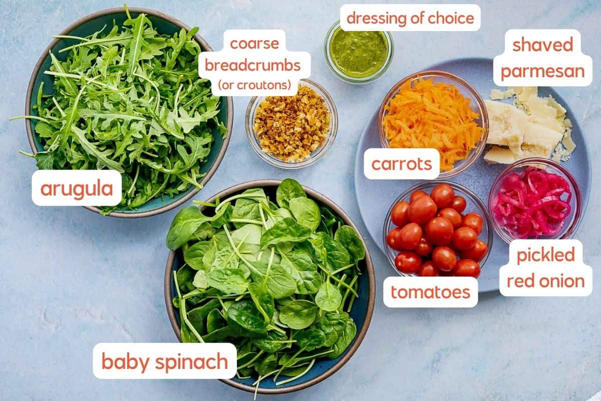 Bowls filled with arugula, baby spinach, shredded carrots, cherry tomatoes, pickled red onions, shaved parmesan, breadcrumbs, and a small bowl of dressing arranged on a blue surface.
