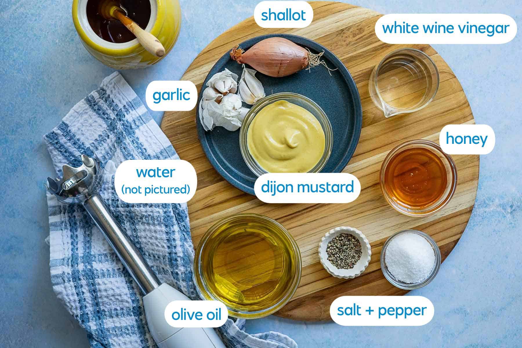 Ingredients for mustard vinaigrette dressing on a wooden board: dijon mustard, honey, white wine vinegar, olive oil, garlic, shallot, salt, pepper, a towel, an immersion blender, and a note indicating water is not pictured.