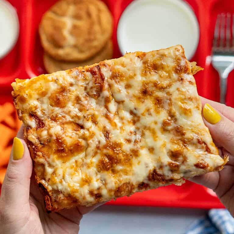 A person with yellow nail polish holds a large, rectangular slice of cheesy pizza over a red cafeteria tray with a biscuit, a fork, and two cups of milk in the background.