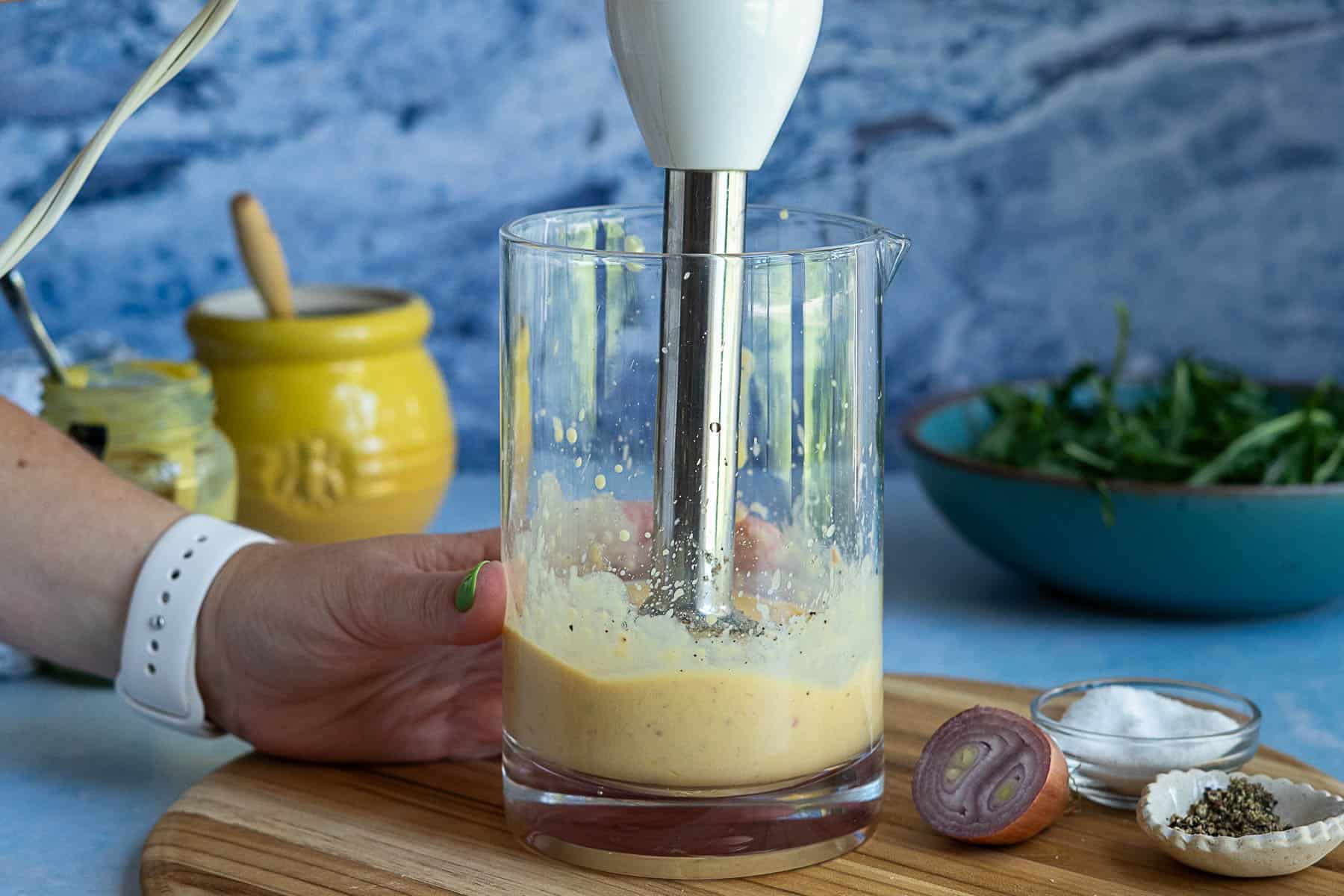 A hand holds a glass container with a stick blender mixing a creamy yellow sauce. Nearby are a sliced shallot, salt, pepper, a yellow jar, and a bowl of greens on a wooden cutting board.