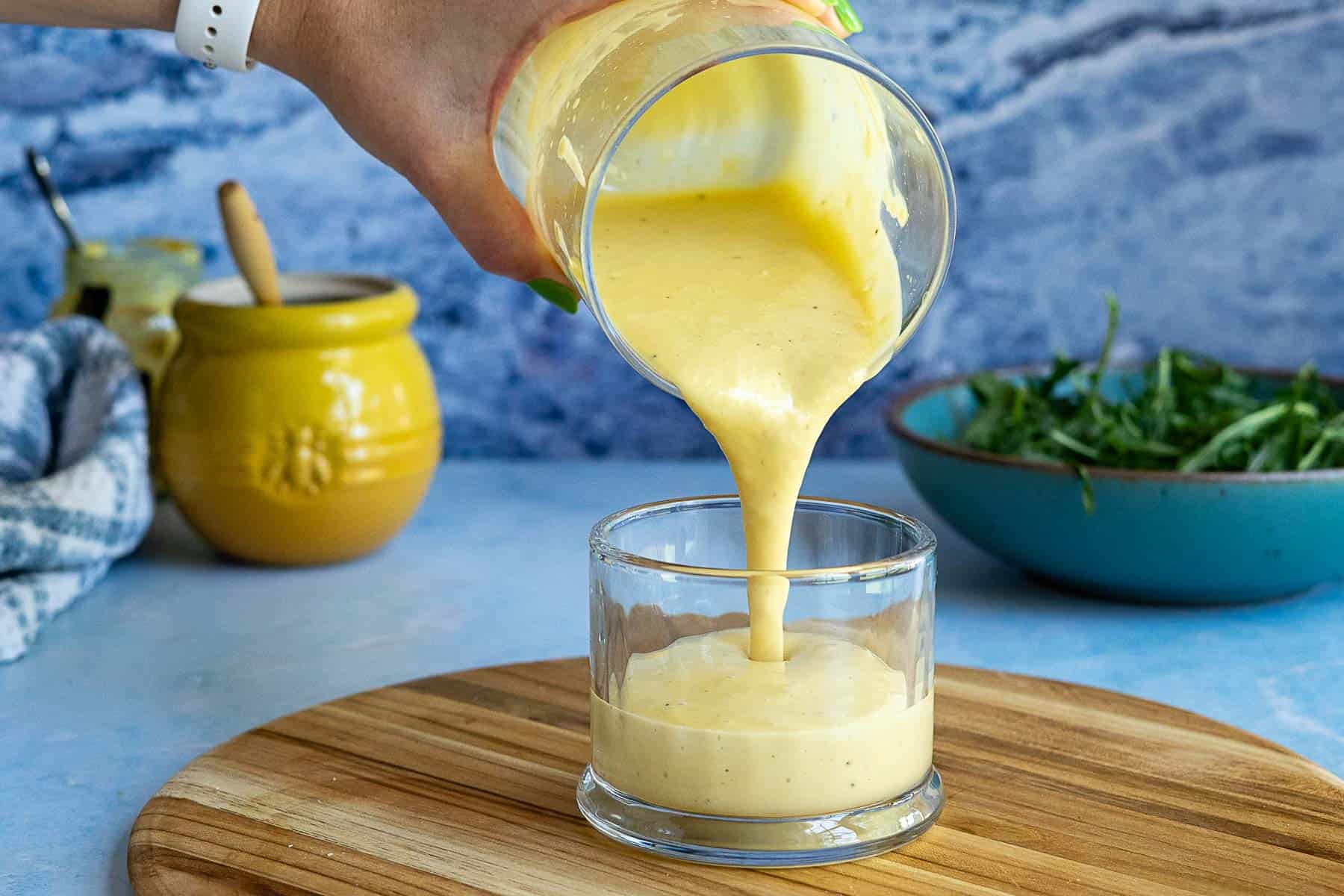 A hand pours creamy Dijon Mustard Vinaigrette from a glass into a small tumbler on a wooden board, with a yellow jar, a bowl of greens, and a blue background in the scene.