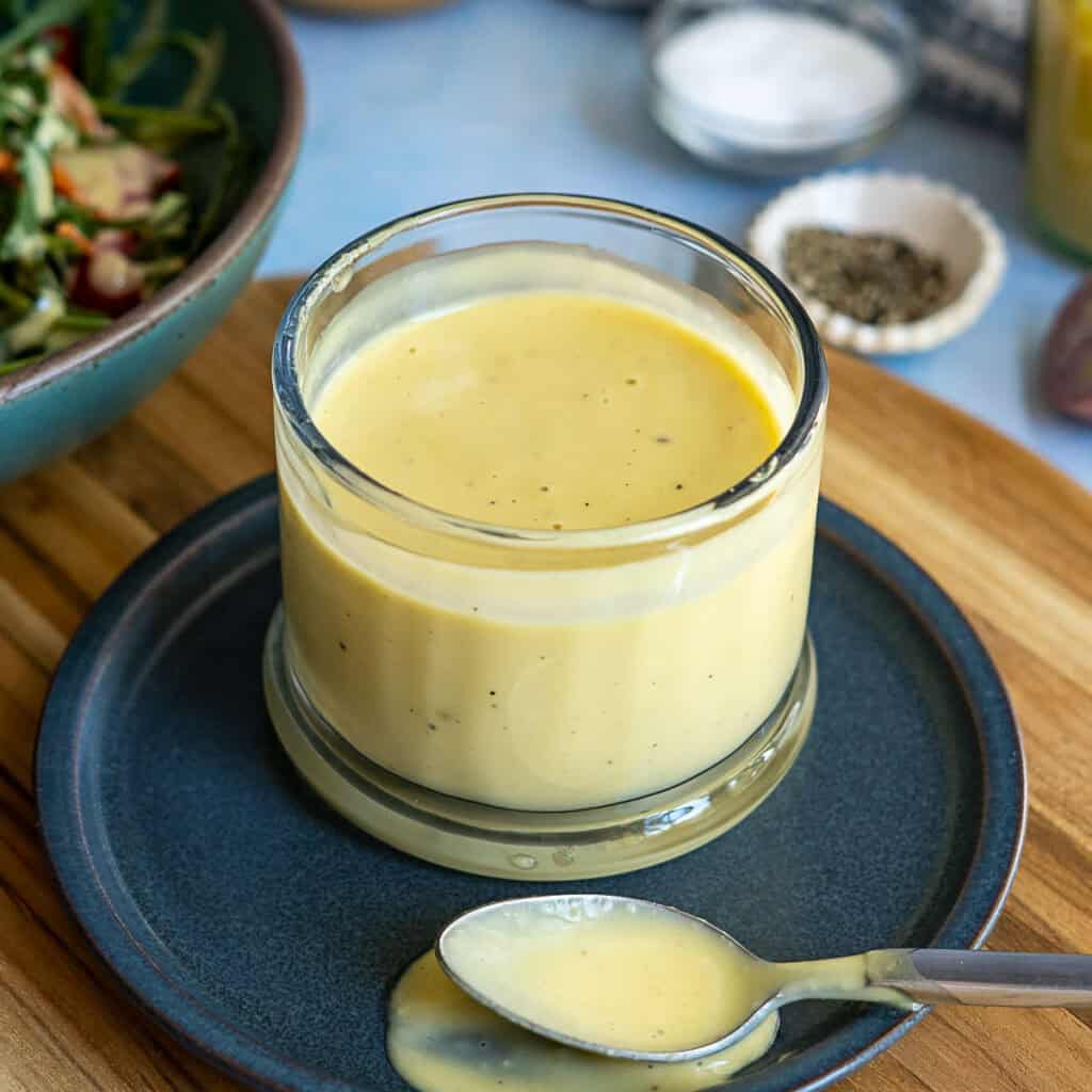 A glass jar filled with creamy dijon mustard salad dressing sits on a dark saucer, with a spoon resting nearby and salad, pepper, and salt visible in the background.