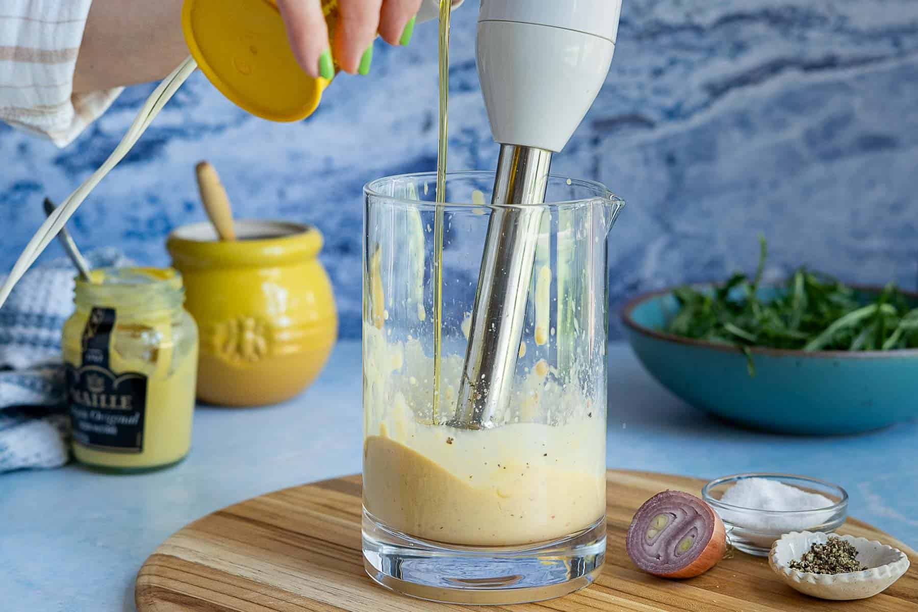 A hand uses an immersion blender to mix ingredients for mustard vinaigrette in a glass jug on a wooden board, with mustard, a shallot, salt, pepper, and a bowl of greens nearby.