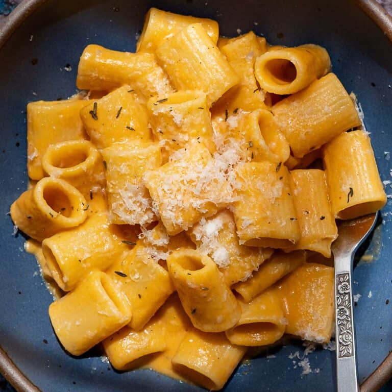 A bowl of rigatoni pasta coated in a creamy orange Butternut Squash Vodka Sauce, topped with grated cheese, with a decorative silver fork resting on the side.